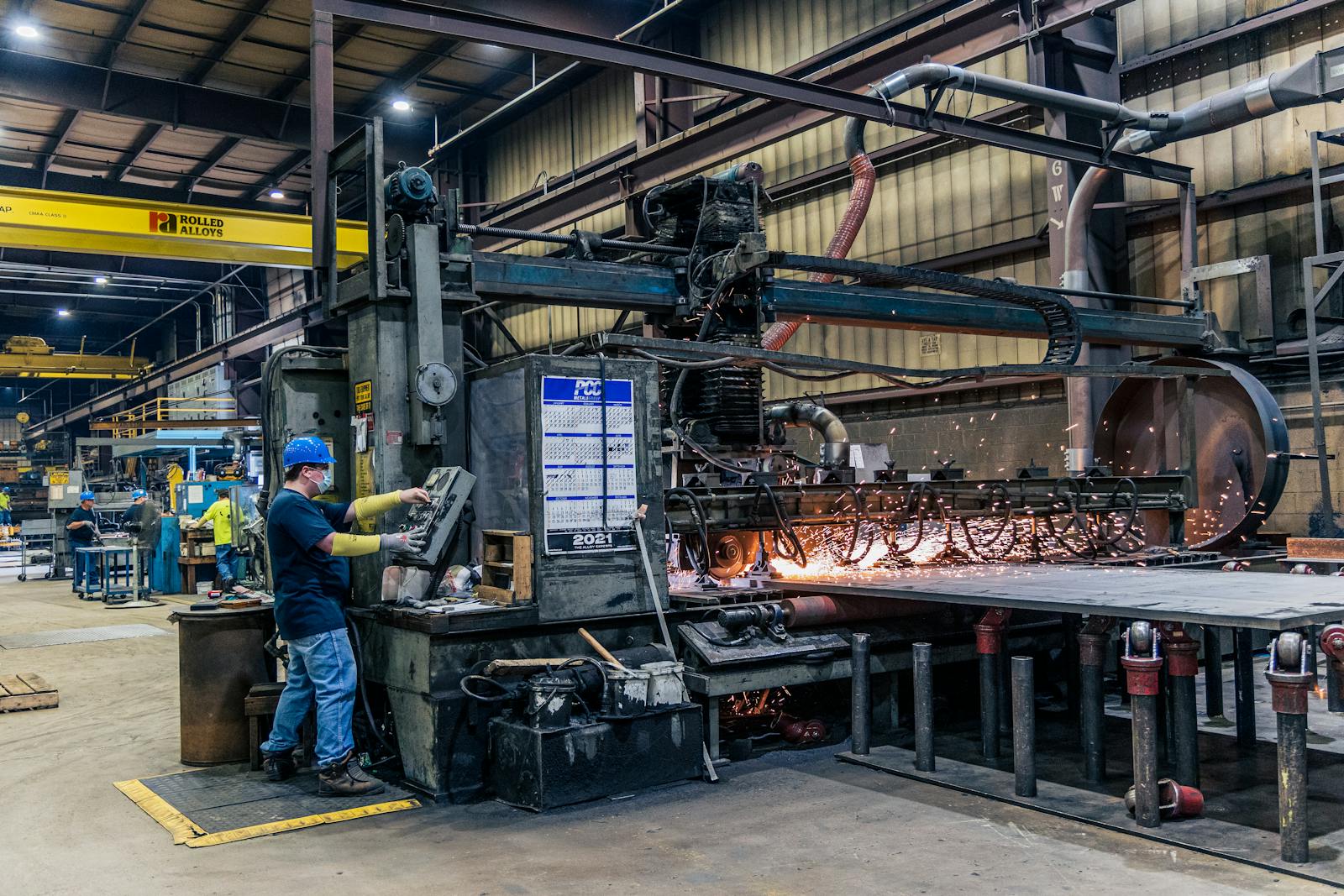 Manufacturing facility floor with heavy equipment, overhead crane, and CNC plasma cutting table producing steel parts